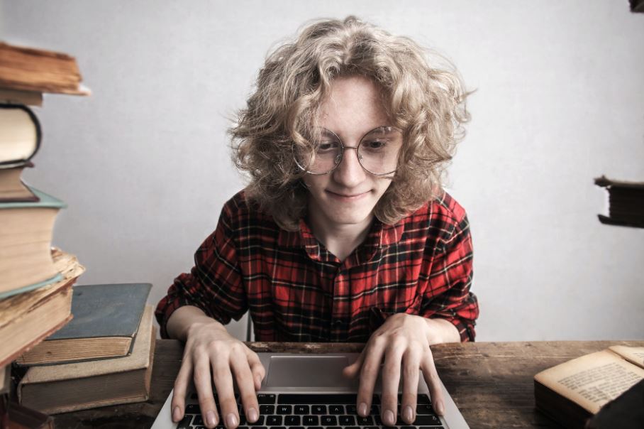 student with glasses using a laptop beside stacks of books